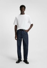 Hvid t-shirt med korte ærmer og præget logo på brystet, kombineret med mørkeblå denim bukser, sikret med et sort bælte og sorte sko.