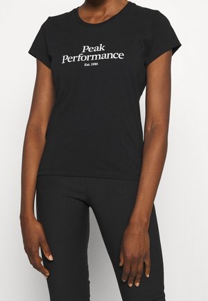 Kobieta ubrana w dopasowaną czarną koszulkę z białym napisem "Peak Performance Est. 1986" oraz czarne spodnie.