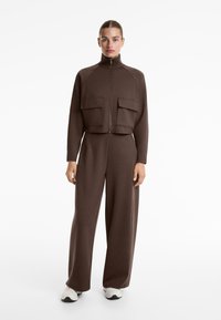 Vrouw die staat, gekleed in een bruin ritsjack met grote voortassen, bijpassende broek met wijde pijpen en witte sneakers op een witte achtergrond.