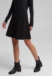 Femme portant un haut noir à manches longues, une jupe trapèze noire avec des petits pois discrets, et des bottines noires, debout sur un fond gris clair.