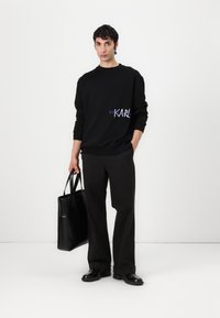 Felpa nera con grafica "KARL" in bianco e viola, indossata con pantaloni e scarpe nere, mentre tiene una borsa tote nera. Stile minimalista.