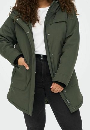 Femme portant un parka à capuche vert olive sur une chemise blanche et un pantalon noir, une main dans une poche avant.