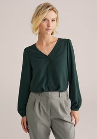 Blusa verde scuro a V con maniche lunghe e a palloncino, caratterizzata da una chiusura con bottoni e una texture morbida, abbinata a pantaloni grigi chiari su misura.
