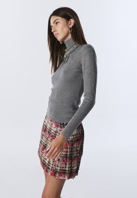 Szary sweter w golf z subtelnym połyskiem, połączony z plisowaną mini spódnicą w kratę w odcieniach czerwieni, żółci i zieleni, akcentowany czarnymi elementami wykończeniowymi.
