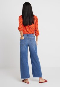 Jeans de mezclilla azules de pierna ancha con cintura alta, bolsillos traseros y dobladillos deshilachados, combinados con una blusa roja de patrón floral brillante.