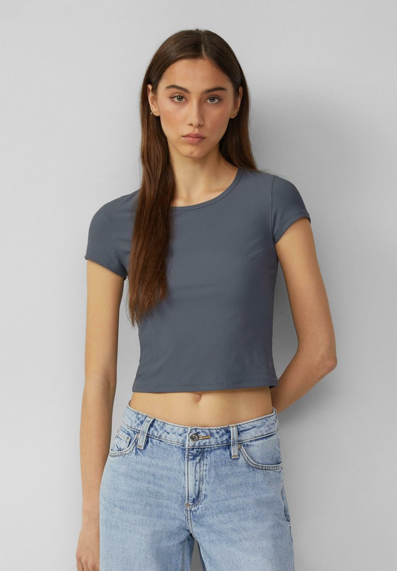 Šedé, krátké cropped tričko s krátkým rukávem z hladkého materiálu, s kulatým výstřihem a minimálními švy, spárováno se světle modrými džíny.