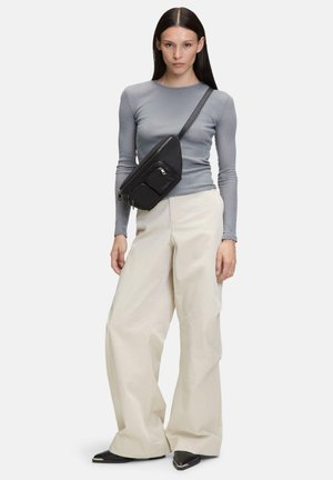 Borsa a tracolla nera con due tasche con zip, posizionata su un modello che indossa un top grigio a maniche lunghe a coste e pantaloni ampi color crema.