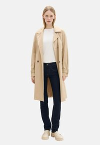 Trenca beige con cierre de botones, llevada sobre una camisa blanca y jeans oscuros. El conjunto se complementa con zapatos de plataforma de color claro.