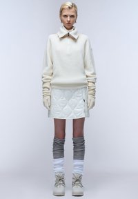 Witte geribbelde trui met hoge kraag en half rits, gecombineerd met een gewatteerde witte rok, grijs en witte kleur legwarmers en neutrale laarzen.