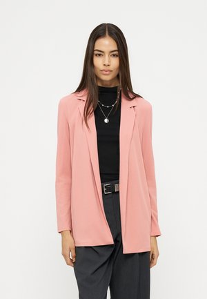 Γυναίκα που φοράει ροζ σακάκι πάνω από μαύρο τοπ με στρωμένα κολιέ και σκούρο γκρι παντελόνι, στέκεται μπροστά σε απλό ανοιχτό φόντο.