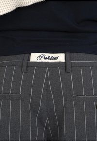 Gros plan d'un pantalon gris à fines rayures avec une étiquette blanche portant la mention « Interdit » sur la ceinture sous une chemise bleu marine foncé.