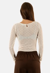 Top a manica lunga in pizzo bianco con motivo geometrico, con scollo rotondo e dettaglio a forma di chiave sul retro. Pantaloni marroni con vita elasticizzata.