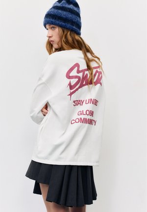 Young woman with long hair wearing a blue striped beanie, white sweatshirt with red text "Stay United Global Community," and a black pleated skirt.