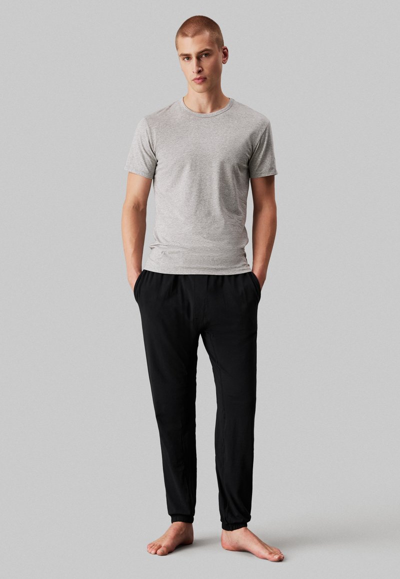 Gray cotton t-shirt and black elastic-waist joggers, featuring side pockets and fitted cuffs, standing barefoot against a neutral background.