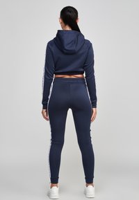 Žena s černým ohonem, oblečená v námořnické modré zkrácené mikině a odpovídajících legínách s bílými bočními pruhy, stojící na šedém pozadí.