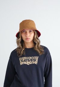 Smeđi šešir s širokim obodom s crvenom donjom stranom, napravljen od tkanine, nosi se s crnim duksericom koja ima uzorak leopard kože i Levi's logo.