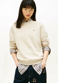 Felpa beige con polsini a costine, indossata sopra una camicia a quadri. Jeans blu scuro con dettagli ricamati. Design semplice e casual.