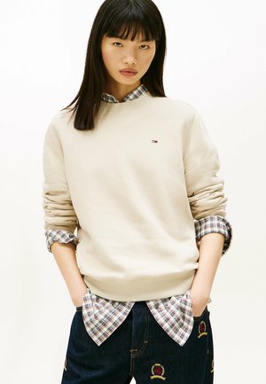 Beige sweatshirt met geribbelde manchetten, gelaagd over een geruit overhemd. Donkerblauwe jeans met geborduurde accenten. Eenvoudig en casual ontwerp.