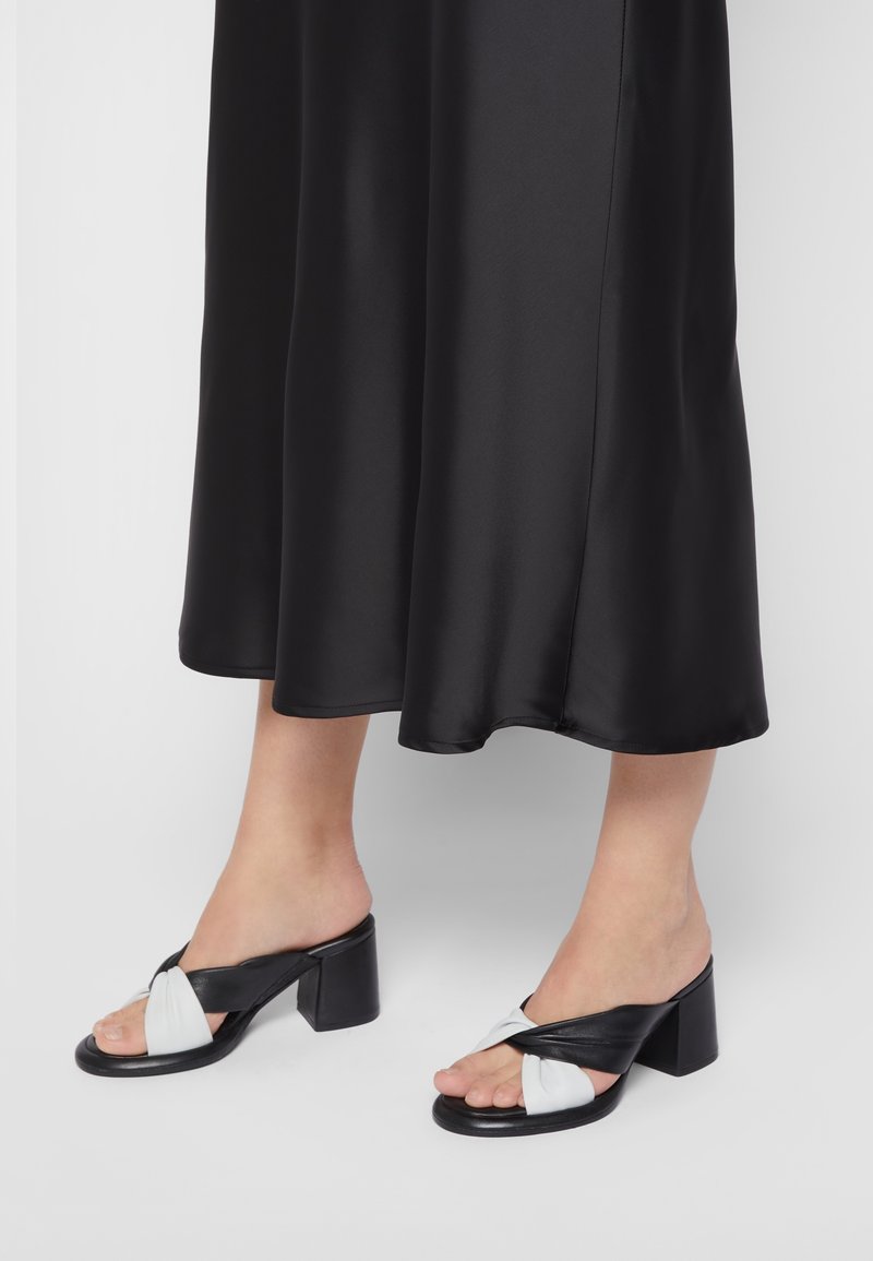 Pieds portant des sandales à talons ouvertes en cuir torsadé noir et blanc, assorties à une jupe midi en satin noir.