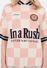 Persoană care poartă o cămașă cu mânecă scurtă, cu model în carouri roz și crem și textul „In a Rush International” și embleme brodate pe piept.