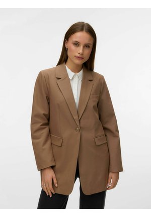 Egyenes barna hajú nő, aki világosbarna, egygombos blézert visel fehér galléros ing felett, egy egyszerű világos háttér előtt áll.