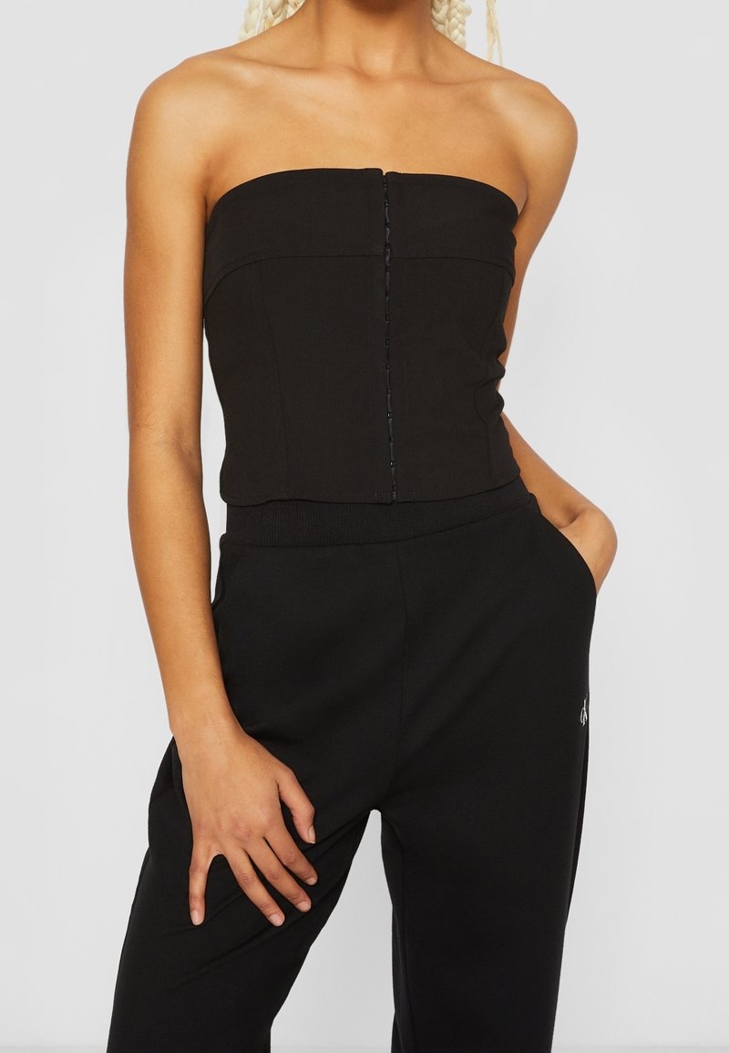 Haut noir sans bretelles au design ajusté, avec un fermeture à boutons sur le devant et un tissu lisse et extensible, associé à un pantalon noir.