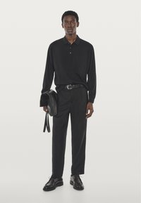 Zwart poloshirt met lange mouwen, zwarte broek en zwarte leren schoenen. Houdt een zwarte leren tas vast, heeft een knoopsluiting en een strak ontwerp.