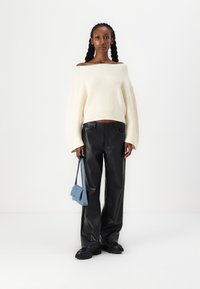 Pull en maille côtelée crème à épaules dénudées, pantalon large en simili cuir noir, et un petit sac à main bleu. Le mannequin se tient devant un fond uni.