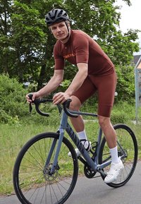Fietser in een bruine, aansluitende jersey en shorts, op een blauwe Canyon-fiets, met een zwarte helm en witte schoenen met sokken.
