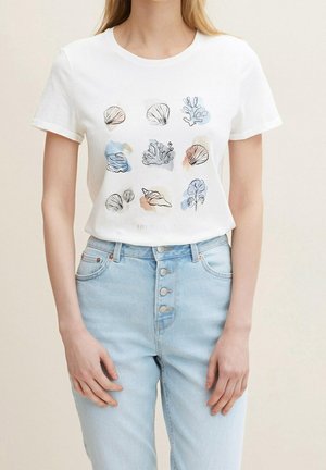 Kobieta ubrana w białą koszulkę z dziewięcioma ilustracjami muszli i koralowców oraz jasnoniebieskie dżinsy z wysokim stanem, z widocznymi czterema guzikami.