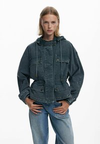 Tume teksaskad, millel on kapuuts, esitaskud, vöökoht on pingutatud ning disain on lõdvestunud. Kangas on tuhm ning tekstureeritud välimusega.