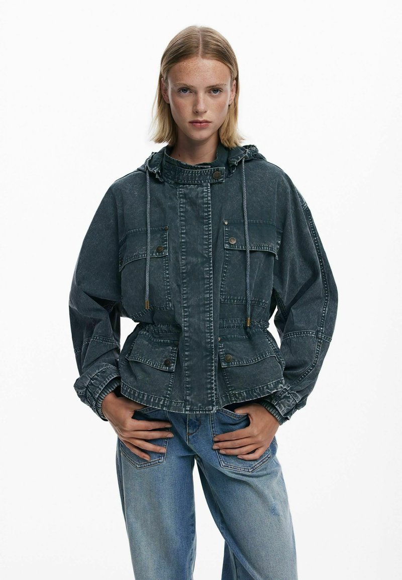 Tume teksaskad, millel on kapuuts, esitaskud, vöökoht on pingutatud ning disain on lõdvestunud. Kangas on tuhm ning tekstureeritud välimusega.