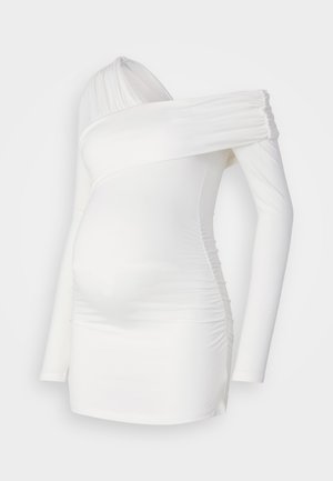 Rochie de maternitate albă cu mâneci lungi, decolteu asimetric, textură pliată pe torace și siluetă ajustată care pune în evidență burtica.