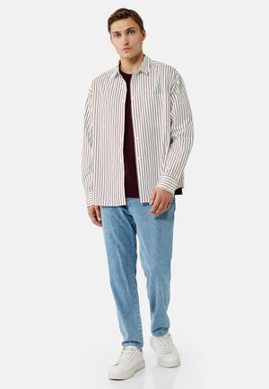 Nuori mies seisoo valkoiset lenkkarit jalassa, vaaleansiniset farkut, viininpunainen paita ja avoin valkoinen pitkähihainen paita pystysuorilla ruskeilla raidoilla.