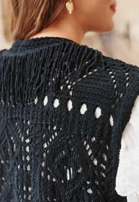 Top in macramè nero con motivi intricati, design a tessitura aperta, frange testurizzate e un aspetto a strati sopra un indumento bianco.