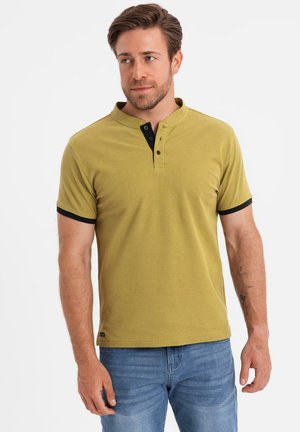 Hombre con cabello corto y castaño que lleva una camiseta henley de manga corta color mostaza con ribete negro y pantalones vaqueros azules, de pie frente a un fondo blanco.