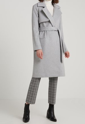 Femme portant un manteau en laine gris clair ceinturé, un col roulé blanc, un pantalon court à carreaux et des bottines noires, debout devant un fond uni.