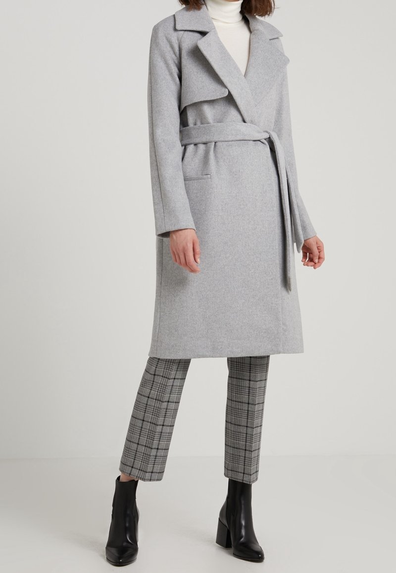 Femme portant un manteau en laine gris clair ceinturé, un col roulé blanc, un pantalon court à carreaux et des bottines noires, debout devant un fond uni.