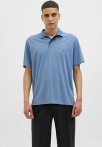 Šviesiai mėlynas polo marškinėliai su trumpomis rankovėmis, sulenkta apykakle ir maža logotipu kairėje krūtinės pusėje. Suderinta su tamsiomis kelnėmis.