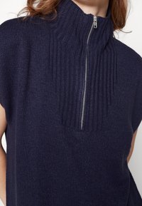 Top senza maniche lavorato a maglia colore blu navy con colletto alto e zip a mezza lunghezza. Presenta un design a costine testurizzato e un tessuto morbido.