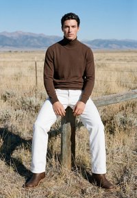 Homme portant un col roulé marron, un pantalon blanc et des bottes marron, assis sur un poteau de clôture en bois dans un champ sec herbeux avec des montagnes en arrière-plan.