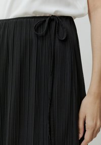 Zwarte geplooide rok met een textuurafwerking, voorzien van een gestrikt tailleband. De stof valt soepel en contrasteert met een lichtgekleurde top.
