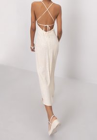 Robe maxi crème avec un motif à pois, design sans dos, bretelles croisées et détail à nouer. Le modèle porte des sandales compensées.