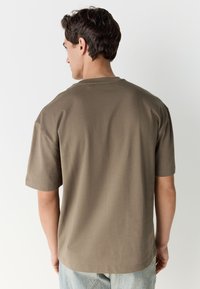 Braunes T-Shirt mit kurzen Ärmeln, aus Baumwolle mit glatter Textur, mit Rundhalsausschnitt und entspanntem Schnitt, Ansicht von hinten.