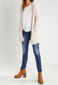 Beige stickad lång cardigan med fickor, lager på en ljusgrå T-shirt. I kombination med blå skinny jeans och bruna klackade ankelskor.