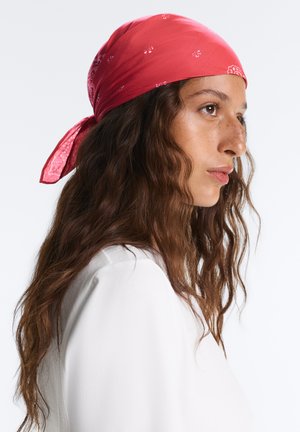 Jeune femme aux longs cheveux bruns ondulés, portant un bandana rouge et une chemise blanche, tournée vers la droite sur un fond clair neutre.