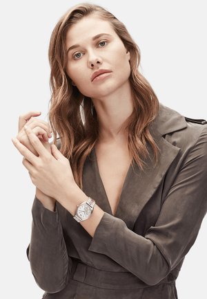 Montre ronde en argent avec un cadran blanc, bracelet en métal et trois boutons de couronne. Le modèle porte un blazer gris. Texture lisse.