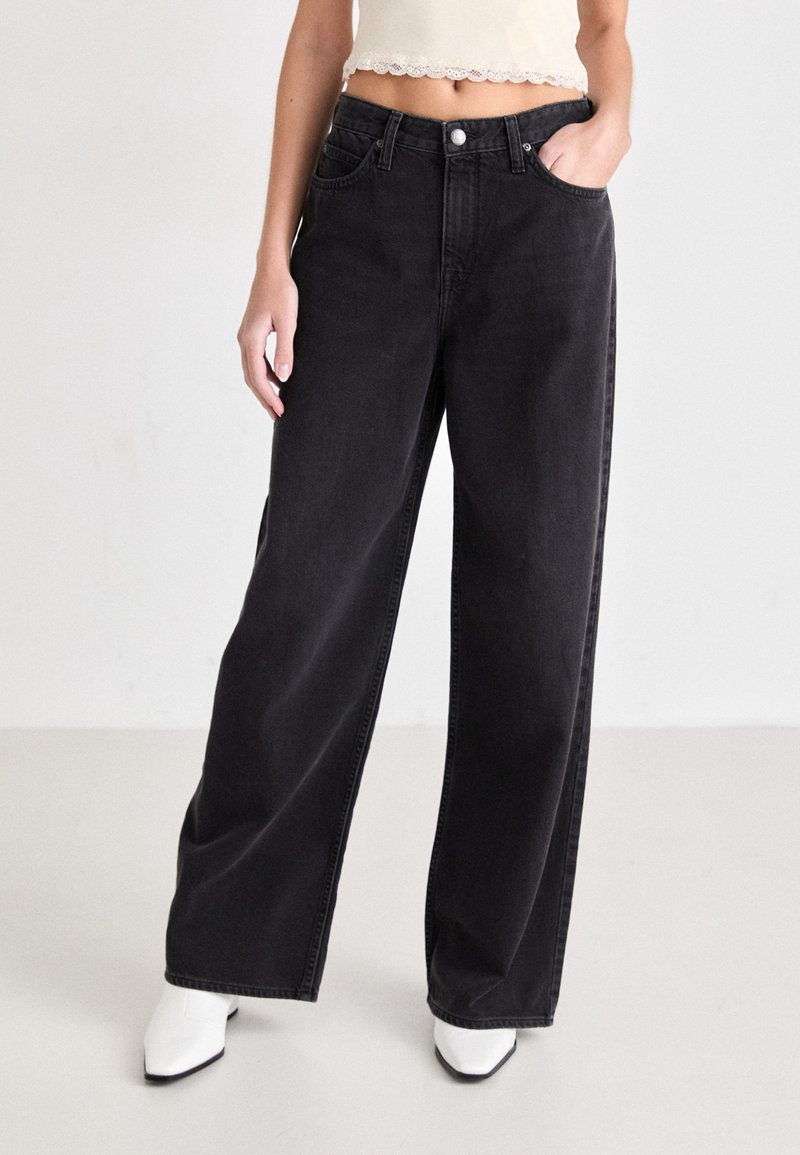 Svarte vide jeans laget av denim, med høy midje, forlommer og knappelukking, stylet med en hvit cropped topp og sko.