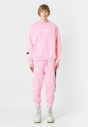 Sudadera de algodón rosa y joggers a juego con acentos blancos y detalles bordados en las rodillas, combinados con botas blancas.