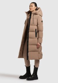 Lange beige pufferjas met een capuchon, voorzien van zwarte ritsen en details. Het model draagt het over aansluitende beige leggingbroeken en zwarte laarzen.